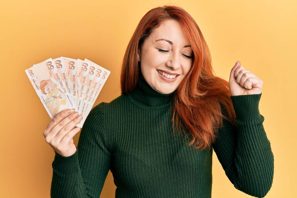 Schöne rothaarige Frau, die 50 türkische Lira-Banknoten in der Hand hält, schreit stolz und feiert Sieg und Erfolg sehr aufgeregt mit erhobenem Arm  - Foto, Bild
