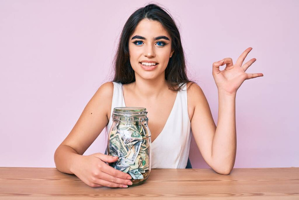 Brünette Teenager Mädchen hält Glas mit Ersparnissen tun ok Zeichen mit den Fingern, lächelt freundlich gestikulierend ausgezeichnetes Symbol  - Foto, Bild
