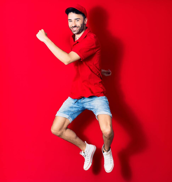 Young handsome deliveryman wearing cap smiling happy. Jumping with smile on face over isolated red background - Φωτογραφία, εικόνα