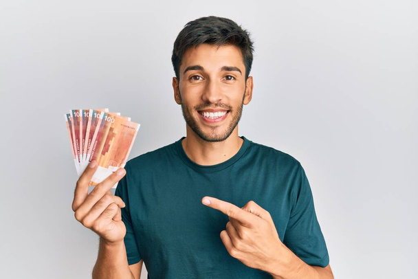 Młody przystojny mężczyzna trzyma 100 norweskich banknotów koronowych uśmiechając się szczęśliwy wskazując ręką i palcem  - Zdjęcie, obraz