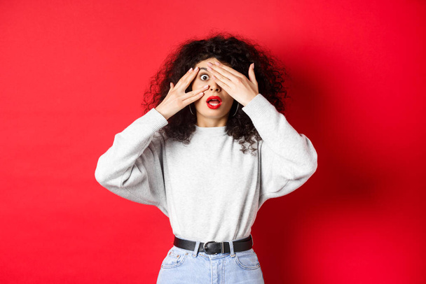 Shocked caucasian girl covering eyes and peek through fingers at something unbelievable, gasping speechless, standing on red background - Zdjęcie, obraz
