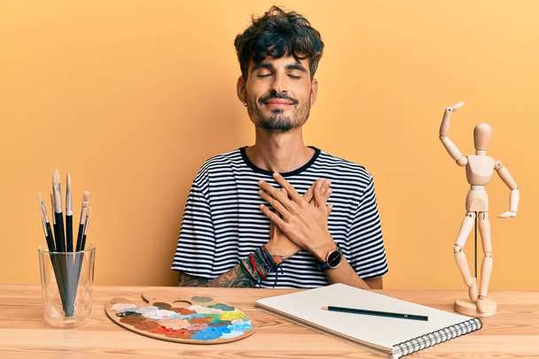 Junger hispanischer Mann, der auf dem Tisch im Kunstatelier sitzt und mit geschlossenen Augen und dankbarer Geste auf dem Gesicht lächelt. Gesundheitskonzept.  - Foto, Bild