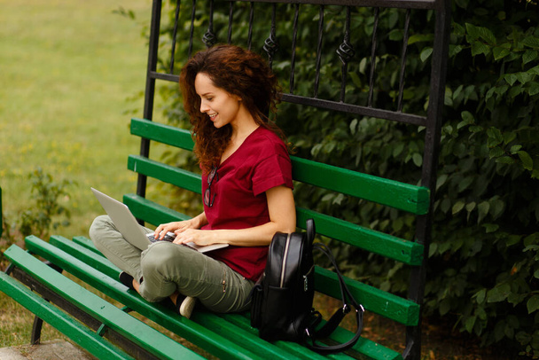 Imagen de una joven rizada sonriente sentada en un banco en el parque y usando una computadora portátil, aislada al aire libre. Vista horizontal. - Foto, Imagen