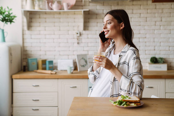 Happy pregnant woman talking on cellphone while having breakfast in cozy kitchen - Foto, Imagen