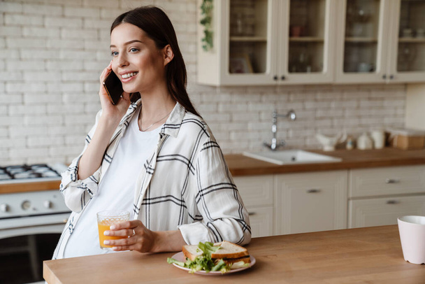 Happy pregnant woman talking on cellphone while having breakfast in cozy kitchen - 写真・画像