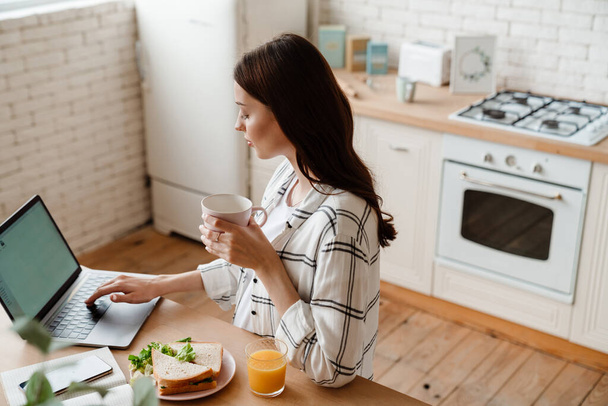 Beautiful focused woman working with laptop while having breakfast in cozy kitchen - Фото, зображення