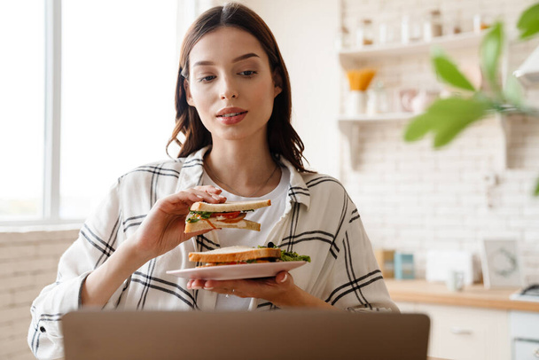 Beautiful focused woman working with laptop while eating sandwich in home kitchen - Foto, Bild
