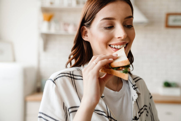 Beautiful happy woman smiling while eating sandwich in home kitchen - 写真・画像