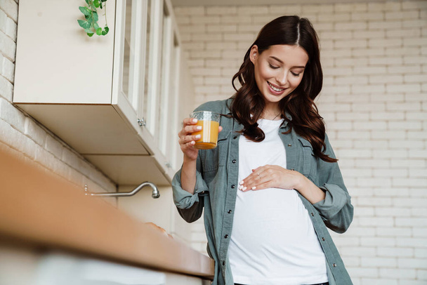 Счастливая очаровательная беременная улыбается и пьет сок на уютной кухне - Фото, изображение