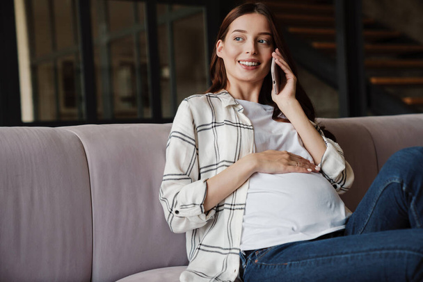 Happy charming pregnant woman talking on mobile phone while sitting on sofa in cozy room - Foto, Bild