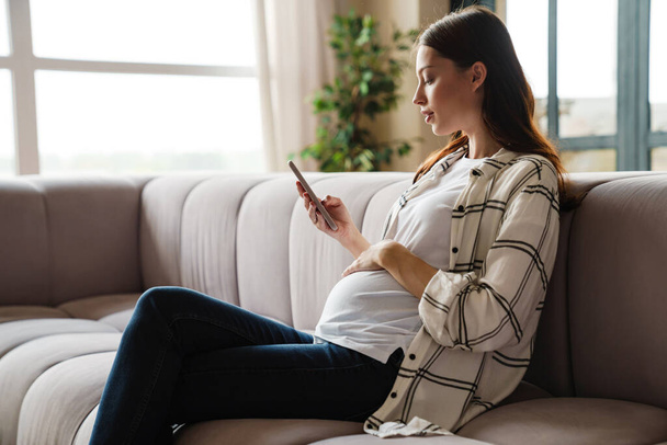 Орієнтована чарівна вагітна жінка використовує мобільний телефон, сидячи на дивані в затишній кімнаті
 - Фото, зображення