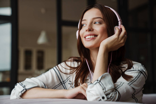 Beautiful happy nice woman listening music with headphones while sitting on couch indoors - Photo, Image