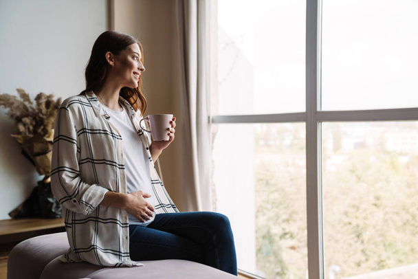 Beautiful happy pregnant woman smiling and drinking coffee while sitting on couch at home - Foto, Bild