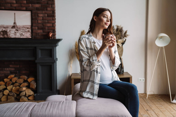 Beautiful happy pregnant woman smiling and drinking coffee while sitting on couch at home - Photo, image