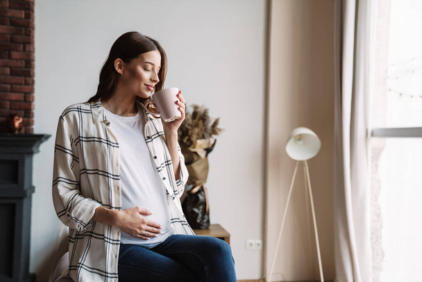 Beautiful happy pregnant woman smiling and drinking coffee while sitting on couch at home - 写真・画像
