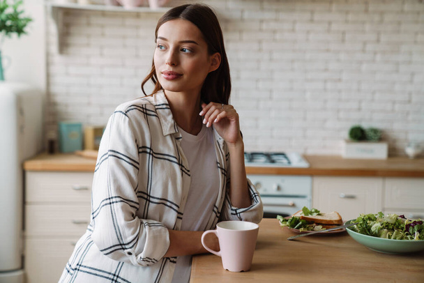 Beautiful pleased woman drinking coffee while having breakfast in cozy kitchen - Photo, Image