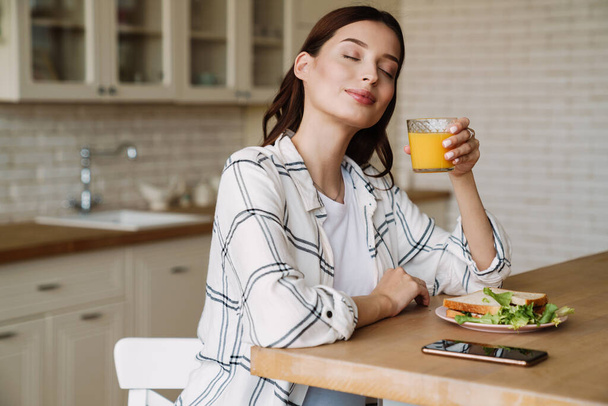 美しい笑顔の女性は居心地の良いキッチンで朝食を持っている間ジュースを飲む - 写真・画像