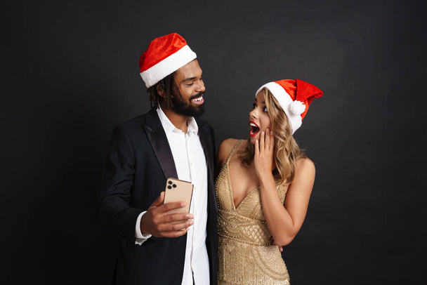 Happy cheerful couple wearing christmas hats using mobile phone isolated over black background - Photo, image