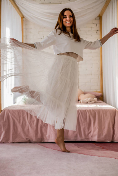 Alegre dama en falda blanca tutu bailando saltando teniendo emoción positiva en el dormitorio loft. Mujer feliz relajante disfrutando de la libertad o fin de semana en el acogedor interior del hogar. Elegante bailarina de moda - Foto, imagen