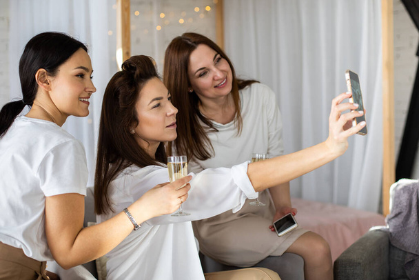 Group of three female friends posing together taking photo use smartphone at home party. Happy women making selfie on mobile phone having fun drinking champagne at home bedroom interior - 写真・画像