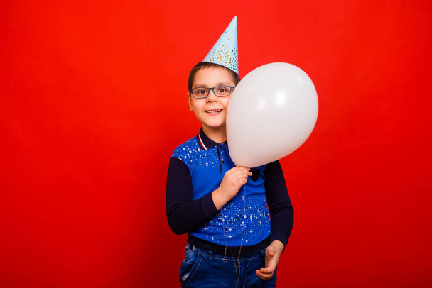 A cheerful boy in a festive cap with a balloon in his hands stands on a red background. - Фото, изображение