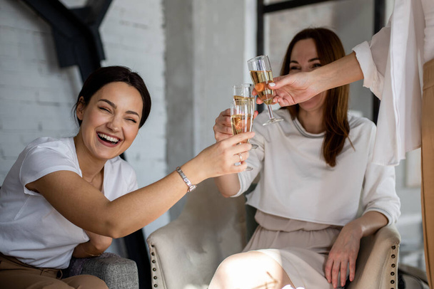 Three happy female friends drinking champagne cheers together laughing having positive emotion low angle. Smiling woman holding glass with cold alcohol beverage rejoicing gossiping enjoying friendship - Photo, Image