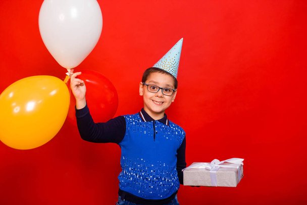 Portrait of a boy in a festive hat with colorful balloons and a box with a bow wrapped in silver paper. A child with a birthday present. Studio photo, standing on a red background. - Фото, зображення