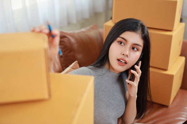 Молода азіатська розумна і активна жінка в повсякденній сукні перевіряє і працює з дому зі смартфоном і онлайн покупкою упаковки коробки замовлення (нова нормальна концепція
) - Фото, зображення