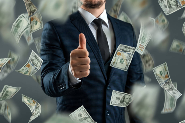 A businessman in a suit shows thumbs up against the background of falling dollars. The concept of investments, dividends, interest, bank deposits - Photo, image