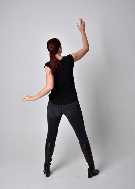 Full length portrait of woman with red hair in a ponytail, wearing black t-shirt and denim pants. Standing pose, hands reaching out with back to the camera the camera, against a  studio background. - Photo, image