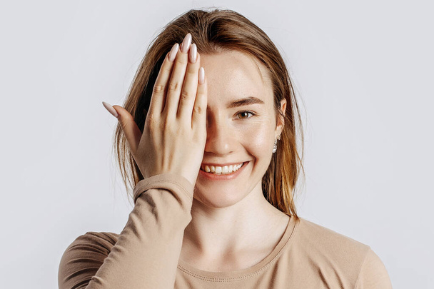 Young beautiful brunette girl smiles and covers half of her face with her hand on a white isolated background - Фото, изображение