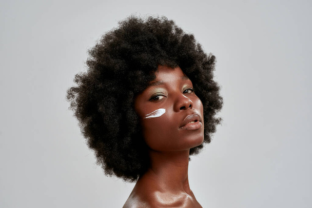 Beauty portrait of young naked african american woman with afro hair looking at camera while posing with cream applied on her face isolated over gray background - Fotografie, Obrázek