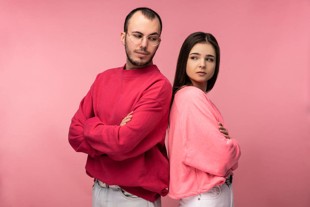 Photo of attractive man wih beard in red clothing and woman in pink stand and pose for camera glare on each other, isolated over pink background - Fotografie, Obrázek