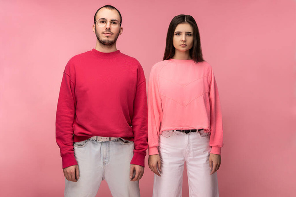 Photo of attractive man wih beard in red clothing and woman in pink stand with serious face, isolated over pink background - Foto, Imagen