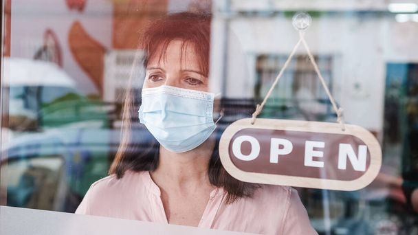 Woman with face mask changing closed to open sign on window looking outside - Photo, Image