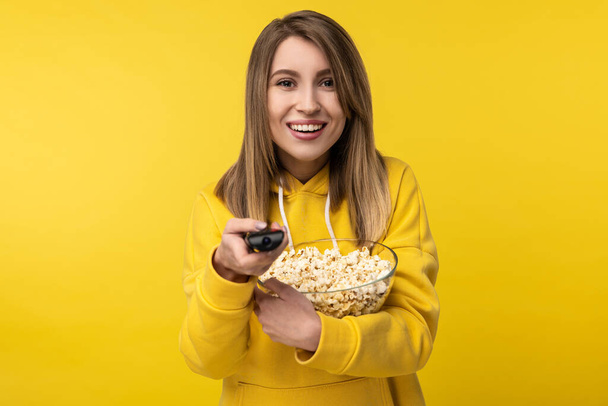 Photo of attractive lady holds tv remote and plate of popcorn, with happy face tries to turn a channel on. Wears casual yellow hoody, isolated yellow color background - Photo, Image