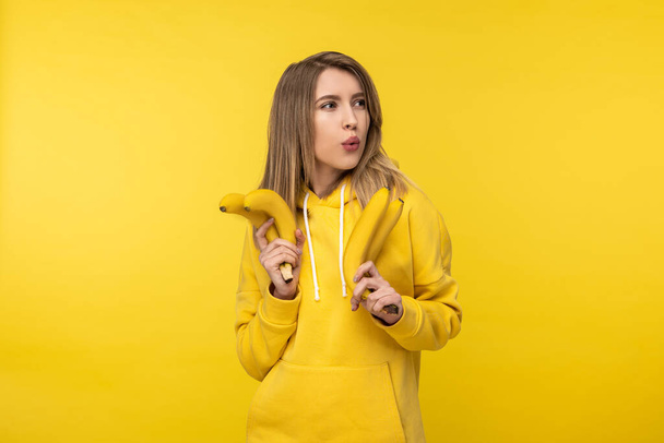 Photo of attractive lady holds bananas and whistles. Wears casual yellow hoody, isolated yellow color background - Foto, afbeelding