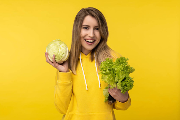 Photo of attractive lady holds cabbage and salad, happy about healthy food. Wears casual yellow hoody, isolated yellow color background - Fotó, kép