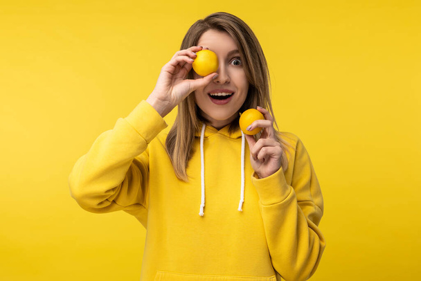 Photo of attractive lady poses in a cute way with lemons, cover one eye up with. Wears casual yellow hoody, isolated yellow color background - Foto, imagen