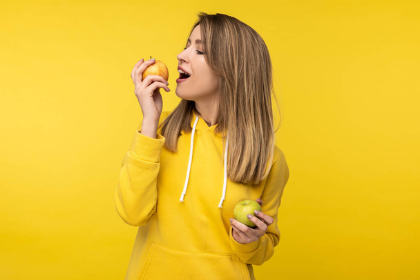 Photo of attractive lady poses in a cute way with apples, tries to bite it. Wears casual yellow hoody, isolated yellow color background - Fotó, kép