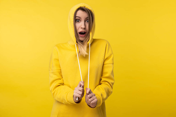 Photo of cute lady pulls on the hood and poses in a funny way. Wears casual yellow hoody, isolated yellow color background - Fotó, kép