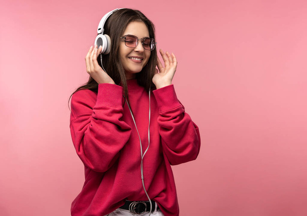 Zdjęcie atrakcyjnej pani w okularach słucha muzyki, uwielbia tańczyć. Nosi luźny różowy sweter białe spodnie odizolowane różowy kolor tła - Zdjęcie, obraz