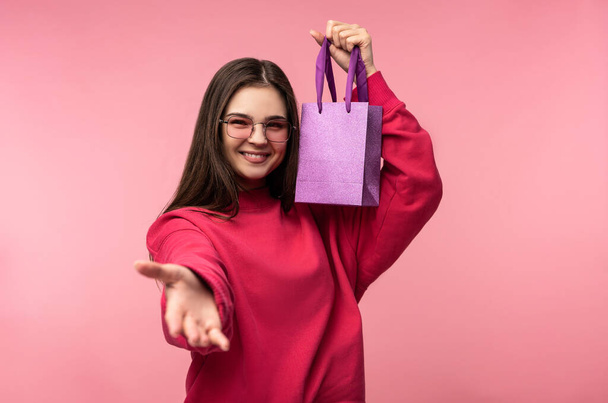 Photo of attractive lady in glasses holds present, pleasant lend a hand, wears casual pink sweater white pants isolated pink color background - Photo, Image
