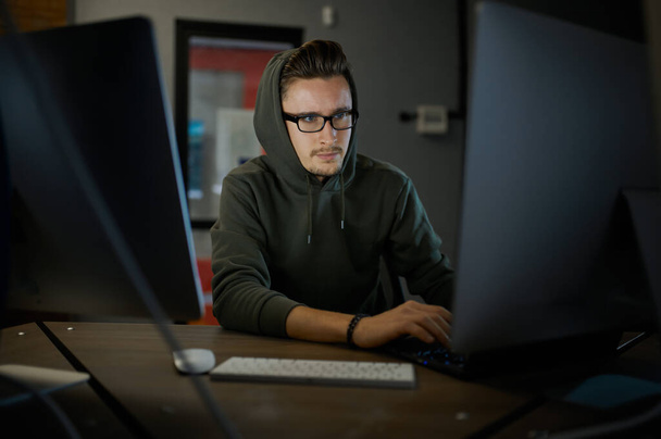 Male internet hacker in hood and glasses sitting at monitors. Illegal web programmer at workplace, criminal occupation. Data hacking, cyber security - Photo, Image
