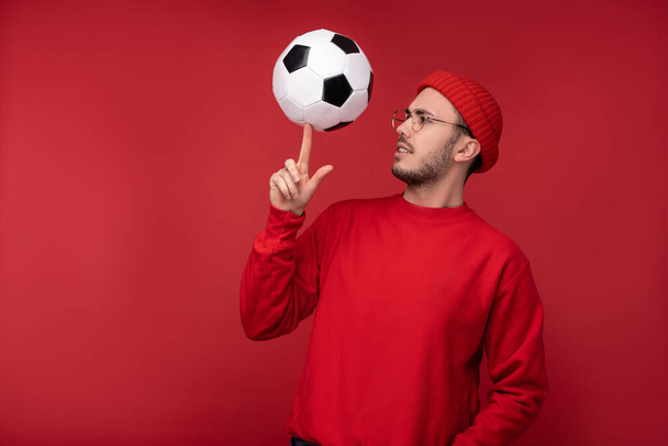 Foto di uomo attraente con la barba in occhiali e vestiti rossi. Felice uomo tiene la palla sul dito indice, isolato su sfondo rosso - Foto, immagini
