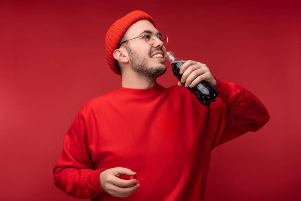 Photo of attractive man with beard in glasses and red clothing. Happy man holds drink, isolated over red background - Фото, изображение