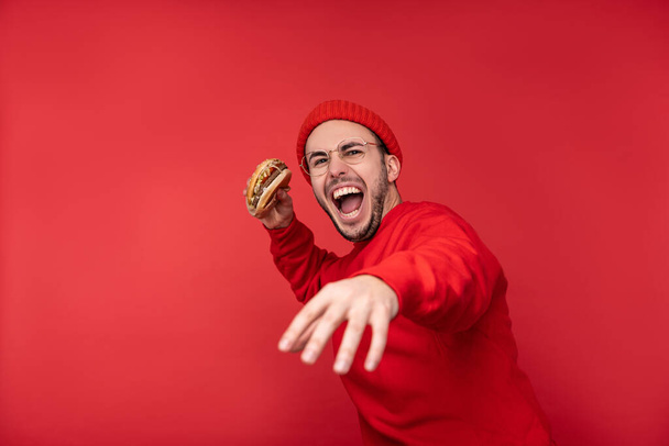 Foto de homem atraente com barba em óculos e roupas vermelhas. Masculino segura hambúrguer e vai jogá-lo, isolado sobre fundo vermelho - Foto, Imagem