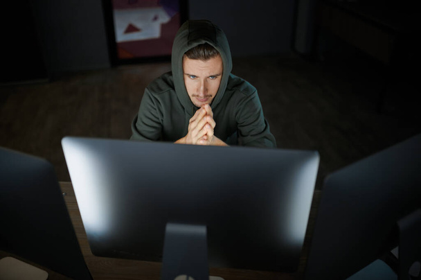 Poważny mężczyzna haker internetowy w kapturze działa na komputerze, widok z przodu. Nielegalny programista w miejscu pracy, okupacja przestępcza. Hakowanie danych, bezpieczeństwo cybernetyczne - Zdjęcie, obraz