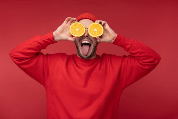 Photo of happy man with beard in red clothing covered eyes with orange fun tongue out, isolated over red background - Photo, image