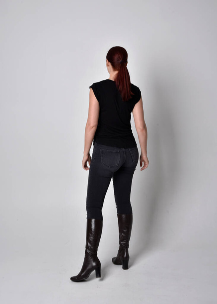 Simple full length portrait of woman with red hair in a ponytail, wearing casual black tshirt and jeans. Standing pose with back to the camera the camera, against a  studio background. - Фото, зображення